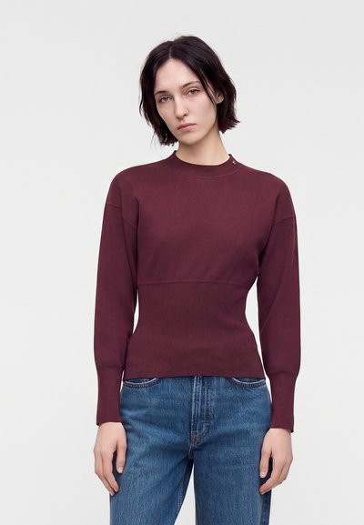 Femme aux cheveux courts et foncés porte un pull bordeaux ajusté à manches longues et un jean bleu, debout devant un fond neutre.