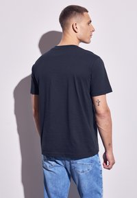 En man som står med ryggen mot kameran, iklädd en svart t-shirt och ljusblå jeans. Korset och små tatueringar är synliga på hans högra arm.