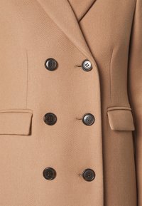 Manteau beige croisé avec six boutons foncés et deux poches à rabat visibles à l'avant.