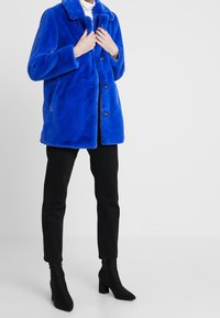 Kungablå fuskpälsjacka med krage, knappstängning och sids fickor, i kombination med svarta smala byxor och klackstövlar.