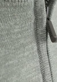 Grijs gebreid stof met een gladde textuur, voorzien van een ritssluiting en een klein metalen label met de gravure "SUITABLE."