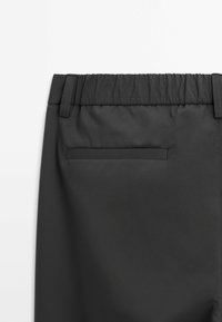 Un pantalon noir à taille élastique présente une poche arrière avec une ouverture rectangulaire, une texture lisse et un design épuré et minimal.