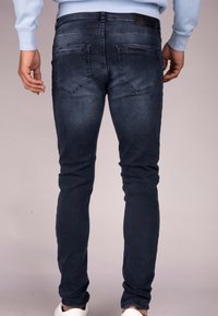 Tmavě modré splývavé džíny s hladkou texturou, obsahující dvě zadní kapsy a koženou nášivku na pase. Viditelné obuv zahrnuje bílé tenisky.