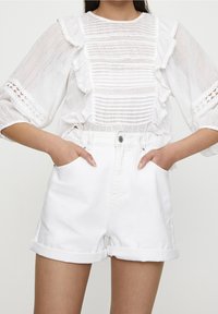 Femme portant un short blanc taille haute avec ourlet roulé et un chemisier blanc transparent avec des détails à volants et des manches trois-quarts.