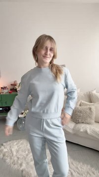 Ung kvinde i lyseblåt afslappet tøj står smilende i en hyggelig stue med en beige sofa, bløde puder og et grønt skab med bøger.