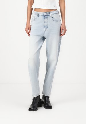 Kvinde iført lyseblå højtaljede jeans, hvid crop top med rib, og sorte ankelstøvler med hæl, stående mod en hvid baggrund.