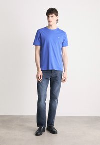 Modrý krátký rukáv tričko s kulatým výstřihem, kombinované s tmavě modrými rovnými džíny a černými botami, stojící na neutrálním pozadí.