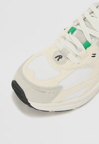 Sneaker con tomaia in tessuto bianco, accenti in suede grigio, suola in gomma nera e dettagli in pizzo verde. Presenta lacci bianchi e un logo rotondo.