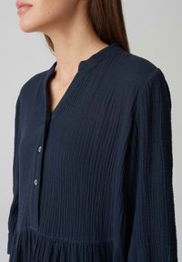 Abito strutturato blu navy con scollo a V, chiusura con bottoni e dettagli plissettati in vita. Presenta maniche lunghe e tessuto leggero.