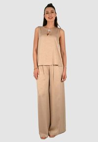 Top smanicato beige con dettaglio ad occhiello, abbinato a pantaloni beige a gamba larga. Texture liscia e design semplice, accessoriato con una spilla floreale.