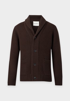 Homme portant un cardigan marron foncé boutonné avec un col châle, une chemise blanche, un jean large bleu clair et des chaussures marron foncé, debout sur fond blanc.