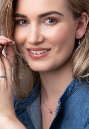 Zilveren ketting met een paarse steen, gecombineerd met zilveren oorbellen en een minimalistische ring. Model in denim jas met licht haar.