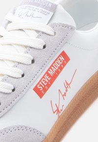 Baskets blanches avec détails en daim gris, lacets blancs, semelle gomme, et logo rouge "Steve Madden Est. 1990" avec signature sur le côté.
