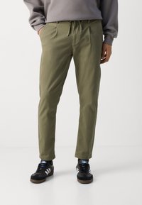 Pantaloni verde măslinie cu talie ajustabilă, croi conic, buzunare laterale și o textură subtilă, asortați cu adidași negri.