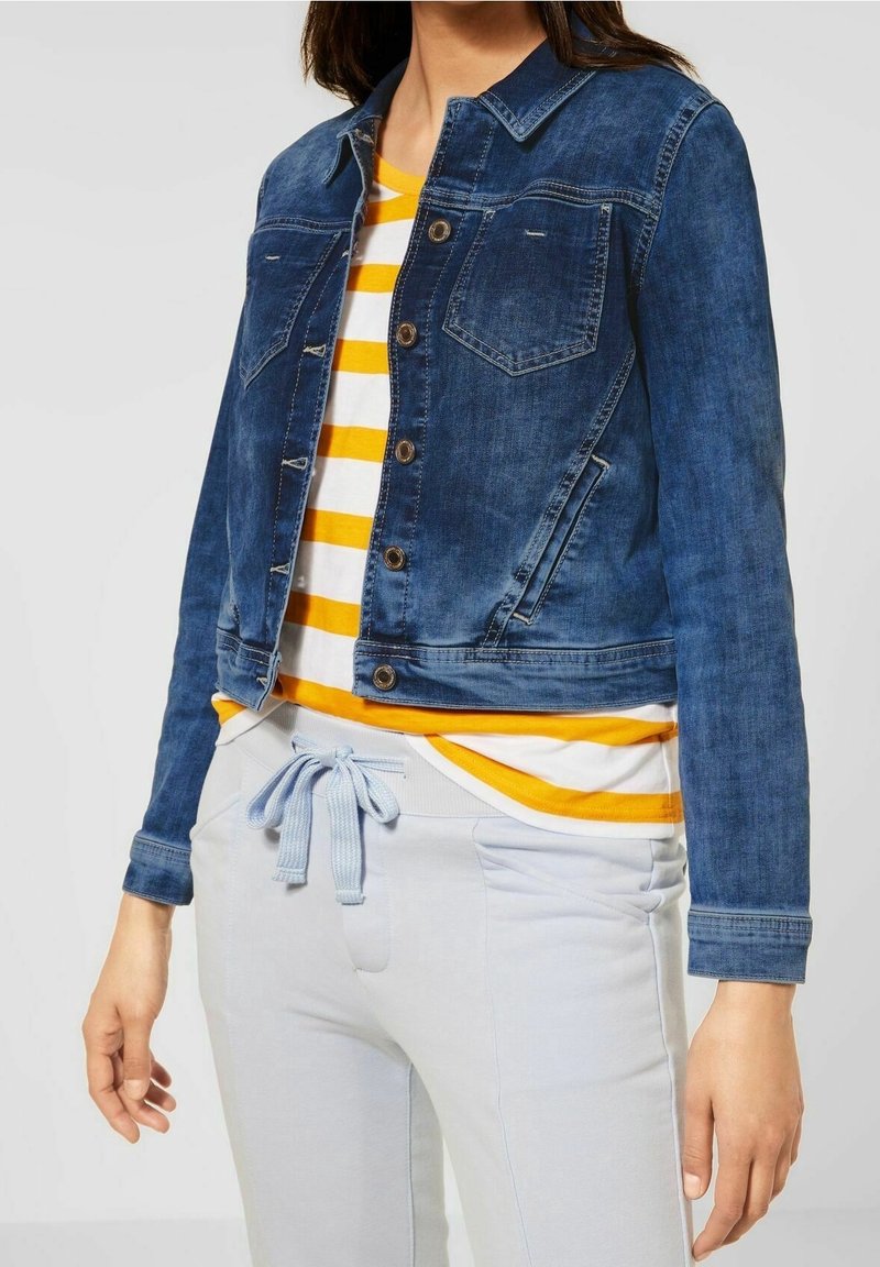 Jeansjacka i mörkblått, med krage, framfickor och knäppning. Parat med en vit och gulrandig topp samt ljusa byxor.