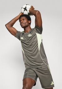 Camiseta atlética gris con detalles en verde claro, de mangas cortas y con logo, combinada con pantalones cortos a juego. El jugador sostiene un balón de fútbol por encima de su cabeza.
