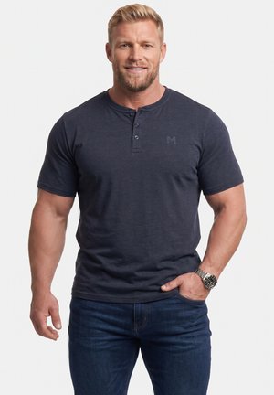 Gespiereerde man met blond haar lacht, draagt een donkerblauw Henley-shirt met korte mouwen, een blauwe spijkerbroek en een zilveren polshorloge.