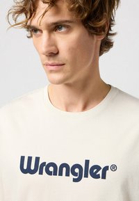 Kermakeltainen puuvillapaita, jossa on suuri, tummansininen "Wrangler" -logo. Klassinen pyöreä kaula-aukko ja lyhyet hihat.