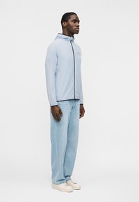 Νεαρός άνδρας που στέκεται, φορώντας ανοιχτό γαλάζιο μπουφάν με κουκούλα και φερμουάρ, φαρδιά ανοιχτό γαλάζια τζιν και λευκά αθλητικά παπούτσια, σε λευκό φόντο.