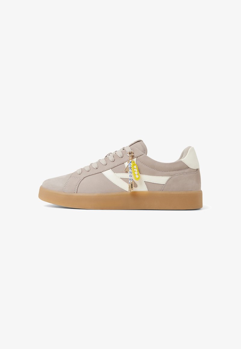 Beige suede sneakers with white accents, flat gum sole, and lace-up design. Features a decorative charm attached to the laces.