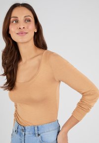 Top a maniche lunghe e aderente in tonalità beige chiaro, realizzato in un tessuto morbido. Presenta un collo a barca e una texture liscia, abbinato a jeans a vita alta.