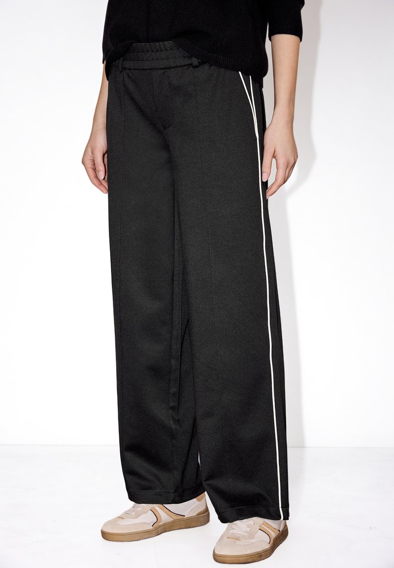 Pantalon noir à jambes larges avec des bandes blanches sur les côtés, porté avec des baskets beige et blanche et un haut noir, modèle debout sur fond blanc.