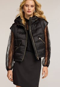 Schwarze gesteppte ärmellose Puffer-Weste mit Kapuze, Reißverschluss, goldener Hardware und gepolsterten Details, kombiniert mit einem langen Oberteil aus Spitze.