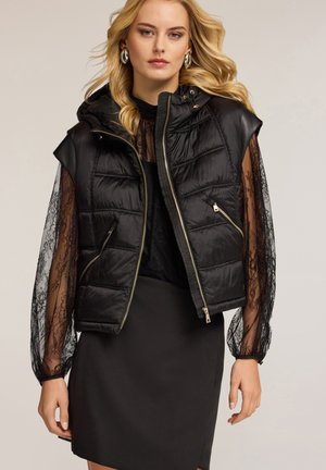 Zwarte gewatteerde mouwloze puffer vest met capuchon, ritssluiting, gouden hardware en gewatteerde details, gecombineerd met een kanten lange mouw top.
