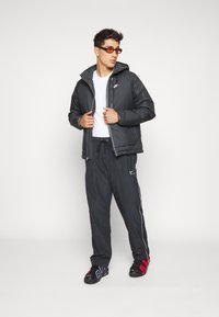 Black puffer jacket with hood, zip closure, and Nike logo; black joggers with white side stripes; black shoes with red and blue accents.