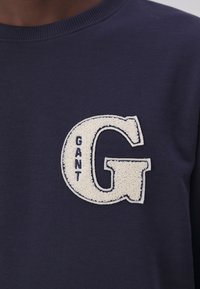 Sweat bleu marine avec une grande lettre blanche texturée « G » avec « GANT » cousu verticalement à l'intérieur de la lettre sur la poitrine.
