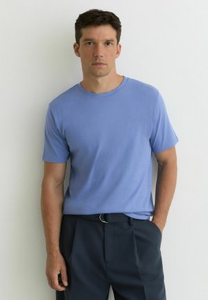 Mand iført en lyseblå t-shirt med korte ærmer og mørke bukser, stående op ad en hvid væg med den ene hånd i lommen.