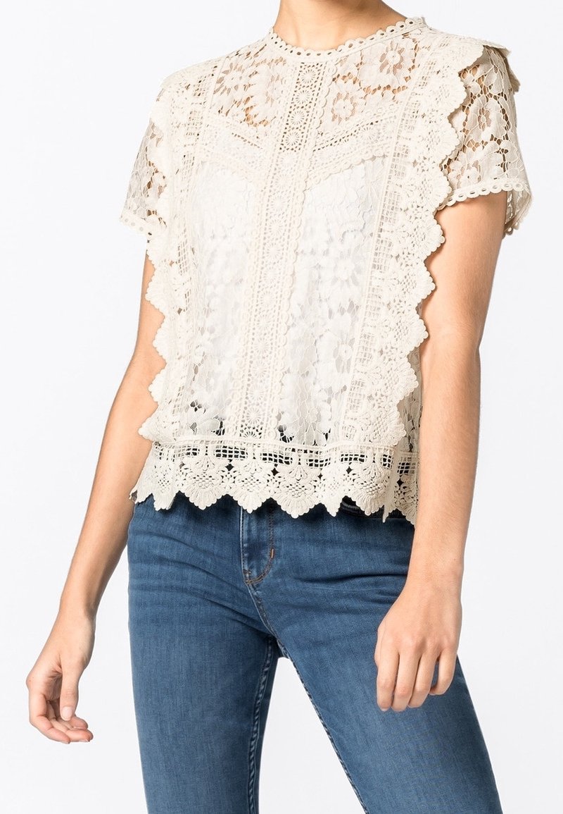 Femme portant un chemisier blanc en dentelle à manches courtes avec des motifs floraux et des bords festonnés, associé à un jean bleu.