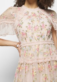 Robe rose pâle avec broderies florales, manches à volants et niveaux ornés de tissu transparent, présentant un design délicat et texturé.