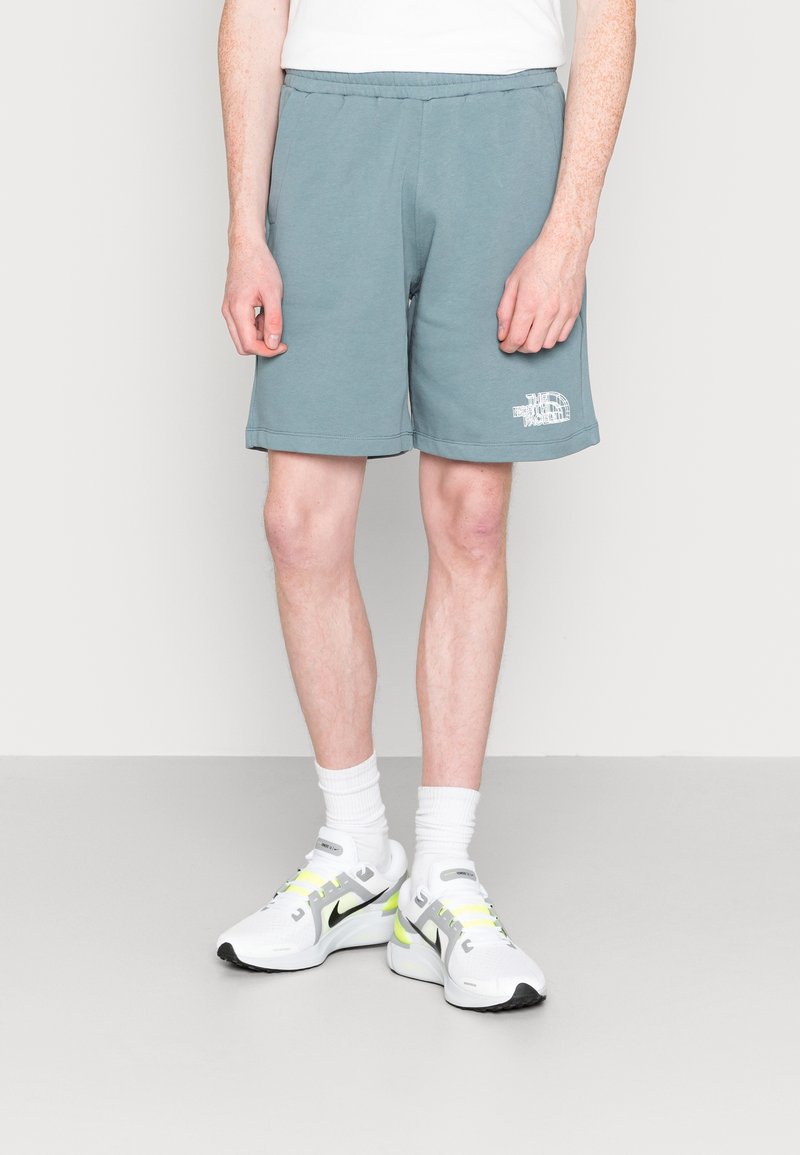 Personne portant un short bleu clair avec un logo blanc, des chaussettes blanches et des baskets blanches avec des accents noirs et jaune néon, debout sur un sol gris.