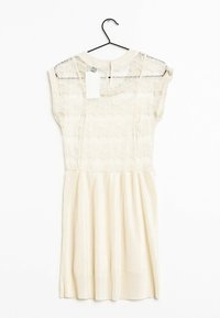 Robe en dentelle avec un haut beige présentant une superposition transparente et une jupe ivoire plissée. Comprend une étiquette, suspendue à un crochet noir sur un fond blanc.