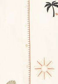 Tissu de couleur crème avec de petits boutons blancs, des lignes cousues marron, un palmier brodé et un motif de rayons de soleil orange.