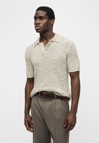 Άνδρας που φοράει κοντομάνικο υφασμένο πλεκτό πόλο πουκάμισο, κρυμμένο μέσα σε καφέ παντελόνι με ψηλή μέση και πλεκτή δερμάτινη ζώνη.
