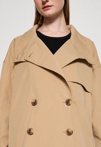 Beżowy płaszcz dwurzędowy z dużymi guzikami, szerokimi klapami i luźnym krojem. Wykonany z gładkiego materiału z widocznymi detalami szwów.