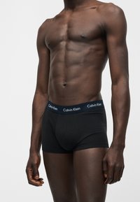 Μυώδες ανδρικό σώμα με σκούρο δέρμα που φοράει μαύρα boxer briefs Calvin Klein με λογότυπο στη μέση, όρθιο σε απλό λευκό φόντο.