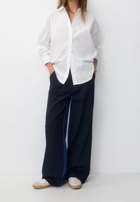 White button-up shirt with long sleeves paired with wide navy pants featuring denim accents and grey sneakers with rubber soles.