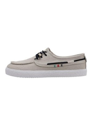 Zapatillas de lona beige con un perfil bajo, acentos de cordones negros y detalles de costura verdes y rojos en el costado. Suela de goma texturizada blanca.
