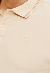 Beżowa koszulka polo wykonana z teksturowanej tkaniny, z zapięciem na guziki i dyskretnym logo. Kołnierz ma kontrastowy pasek.