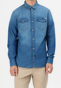 Camicia in denim blu con maniche lunghe, colletto a punta, due tasche sul petto e bottoni in metallo bianco. Texture liscia, design casual.