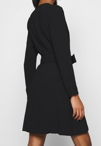 Robe noire ceinturée avec une taille cintrée, des manches longues et une jupe évasée. Tissu lisse avec des plis subtils à l'arrière pour plus de détails.