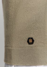 Beige gebreide trui met geribbelde zoom, voorzien van een zwart hexagonaal logo-opdruk met drie horizontale strepen in goud. Zachte textuur en aansluitend ontwerp.