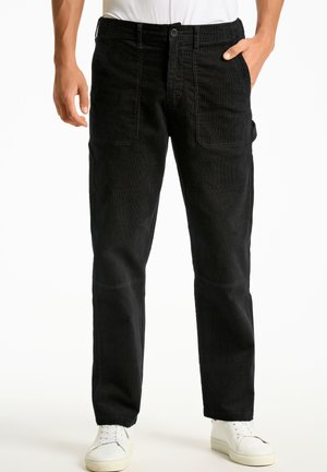 Pantaloni di velluto a coste neri con vestibilità dritta, dotati di tasche laterali e chiusura a bottone, abbinati a sneakers bianche.