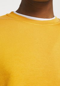 Gele crewneck trui met een gestructureerde stof, voorzien van een witte binnenkraag. Close-up weergave toont de geribbelde halslijn en het gladde lichaam.