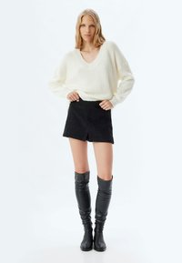 Cremfarbener V-Ausschnitt-Pullover, schwarzer, strukturierten Minirock mit Schlitzen, knielange schwarze Stiefel. Weicher Stoffmix, entspannte Passform und glatte Textur.
