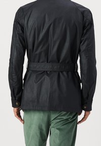 Musta vedenpitävä takki, jossa on vyötäröllä vyö, sileä pinta, pitkät hihat napillisine kalvosimineen ja hieman kaareva helma.