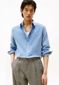Lichtblauw overhemd met opgerolde mouwen, gedragen over een wit shirt, gecombineerd met bruin geruite high-waisted broek.